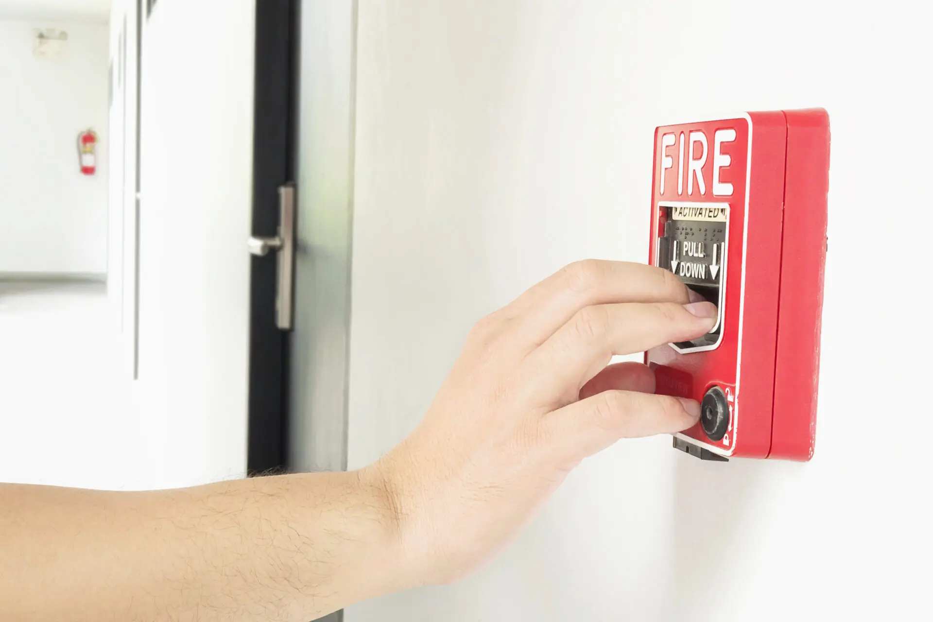 Eine Hand betätigt den roten manuellen Brandmelder mit großer FIRE-Aufschrift und zieht den Auslöser nach unten.