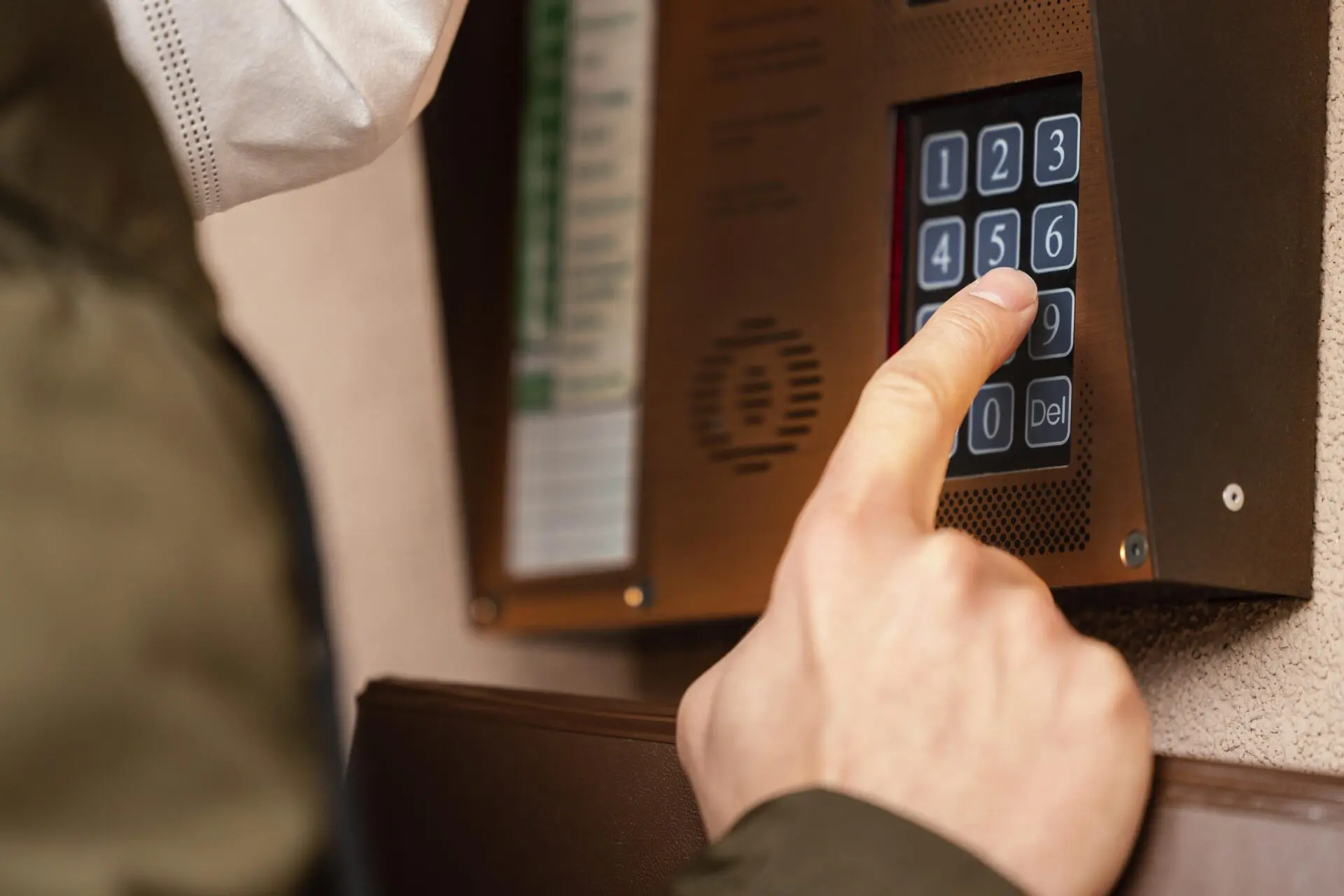 Ein Finger drückt eine Taste der numerischen Tastatur an einer braunen Türsprechanlage mit sichtbaren Sprechöffnungen.